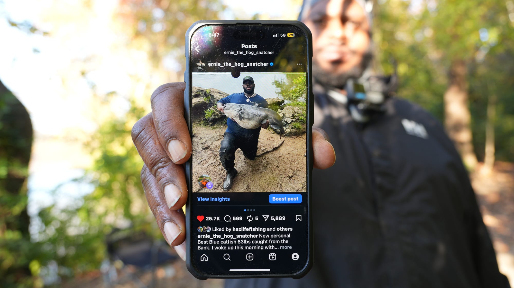 Ernie the Hog Snatcher holds up a phone showing one of his social media posts — catching a "new personal best" 63-pound blue catfish.