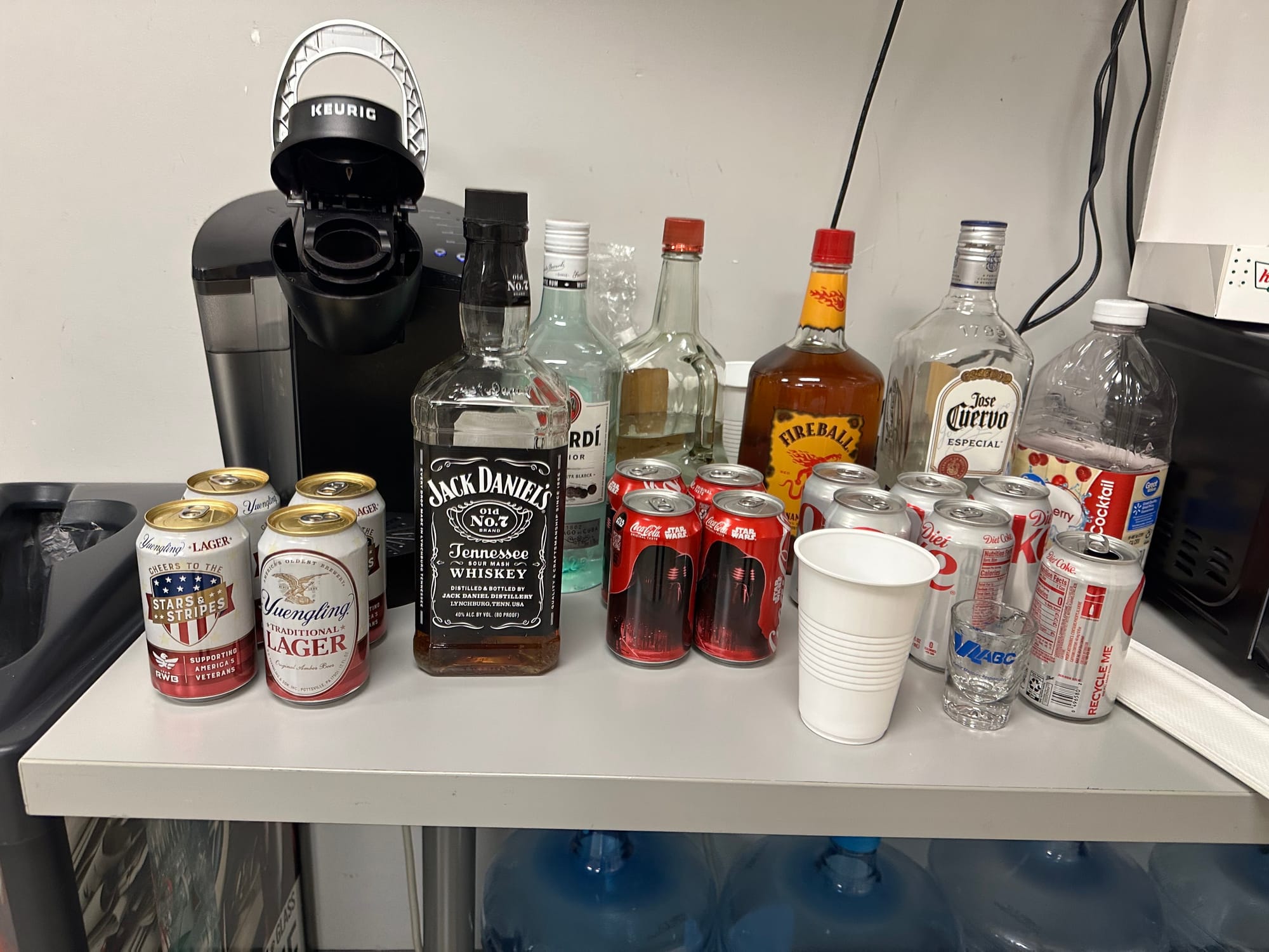 An office table with a Keurig and various types of alcohol on it:&nbsp;Beers, Jack Daniels, Bacardi, Fireball, and Jose Cuervo.