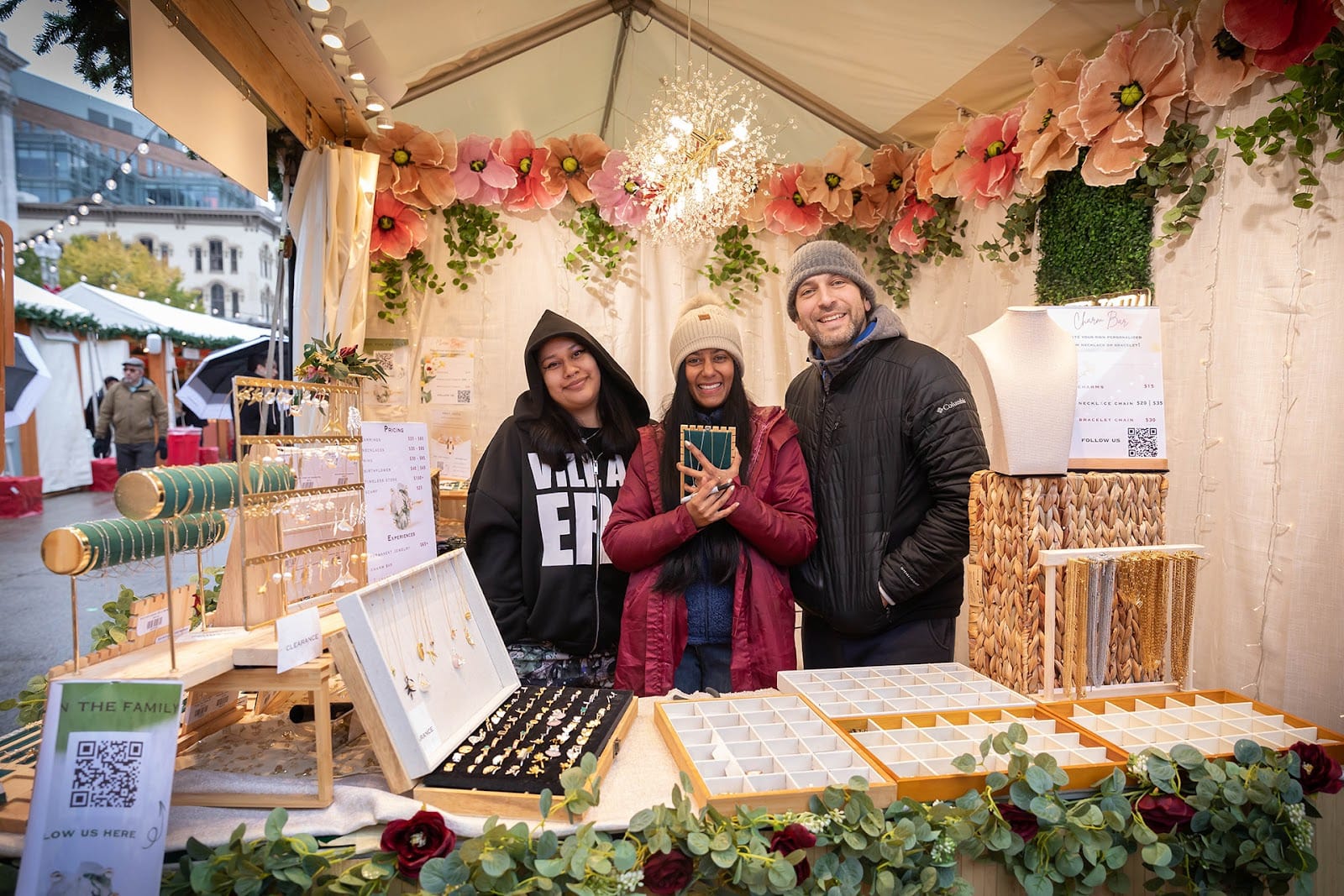 Three people smile while selling jewelry at a holiday market in a cloth booth filled with fake flowers and greenery.