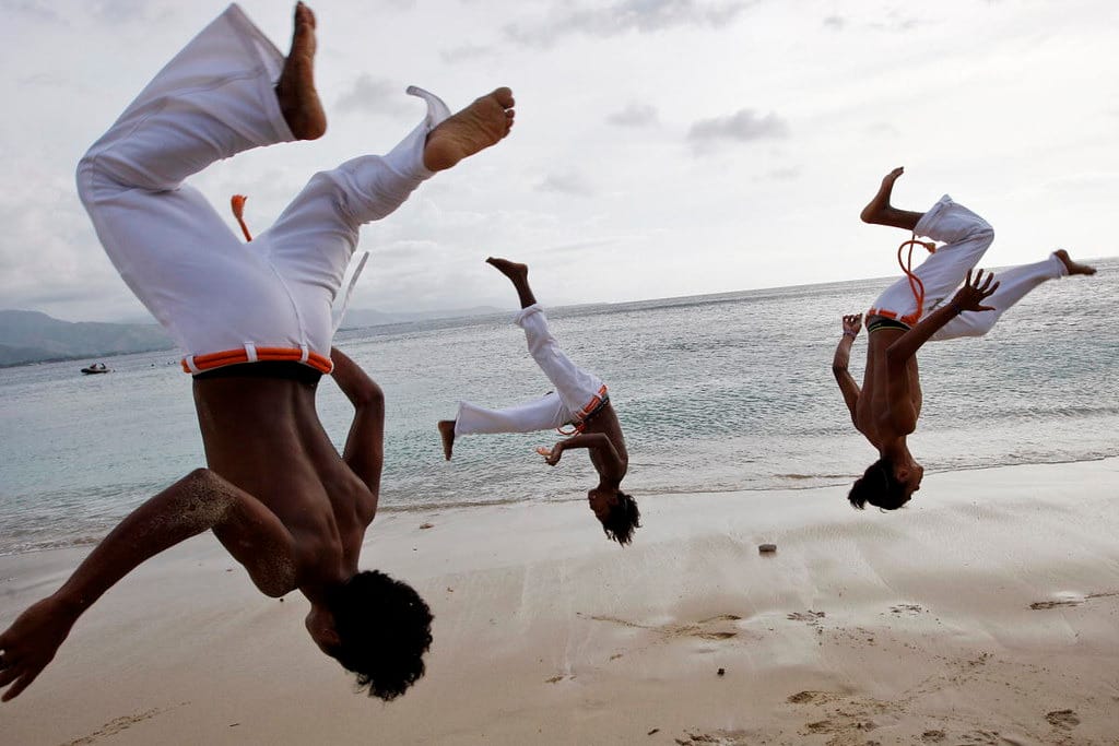 Three people flip upside down on a beach, wearing matching white pants.