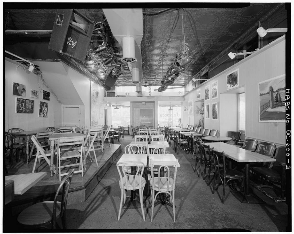 A black and white photo shows the empty inside of d.c. space —&nbsp;featuring tables and chairs arranged around a large room. 