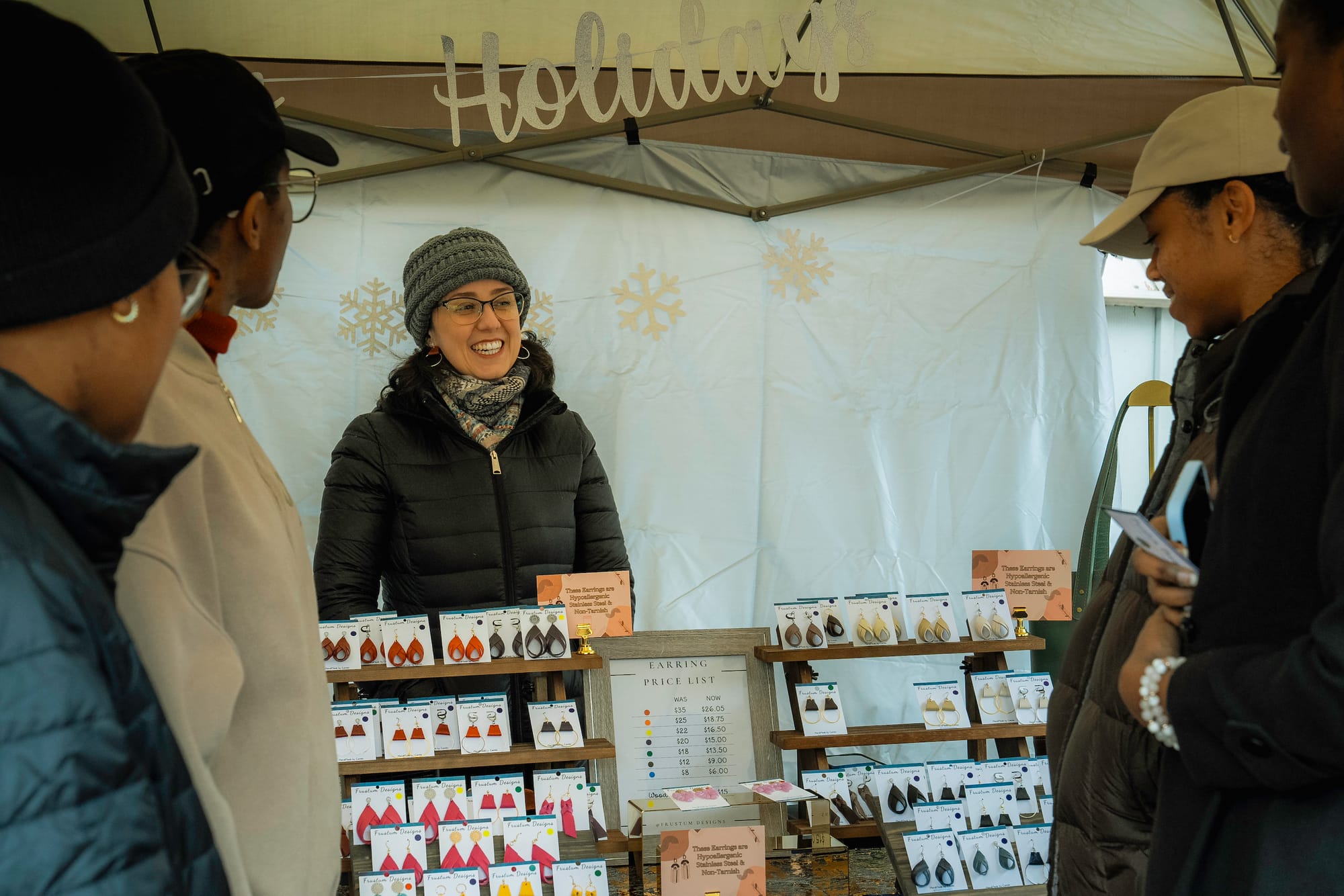 Four people look at earrings sold by a vendor an outdoor holiday market.
