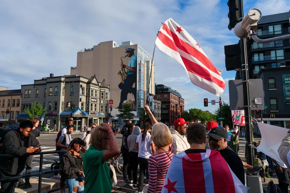 A crowd standing at an intersction, a person draped in a DC flag and another holding one up high.