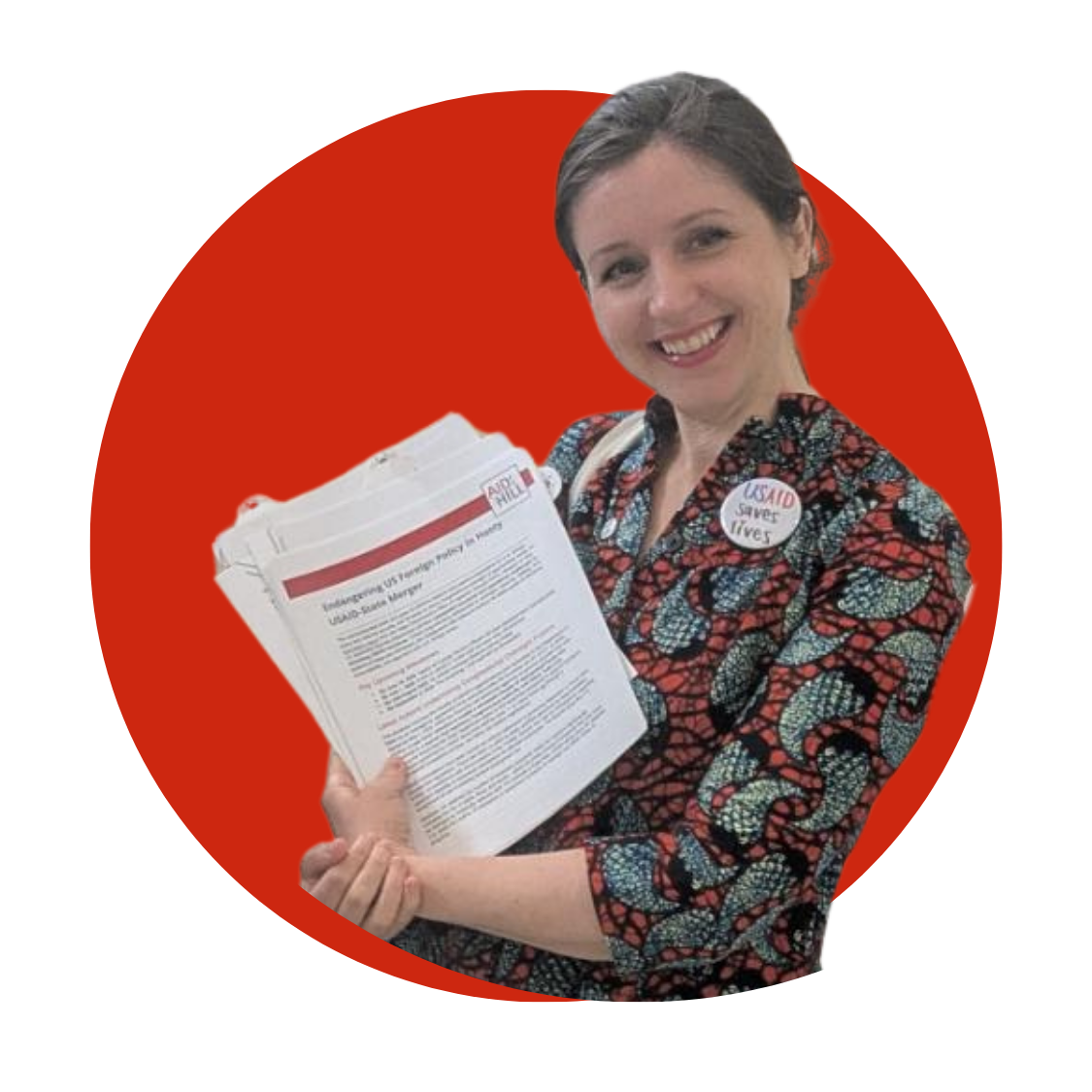 A portrait of Juliane Weis against a red background. She's holding a stack of papers and wearing a patterned shirt and button that says "USAID saves lives"