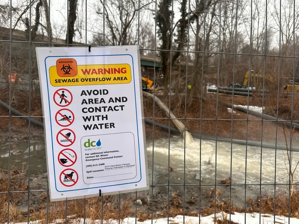 A sign on a fence A sign reads "warning sewage overflow area" in front of a pipe spilling water into a canal. 