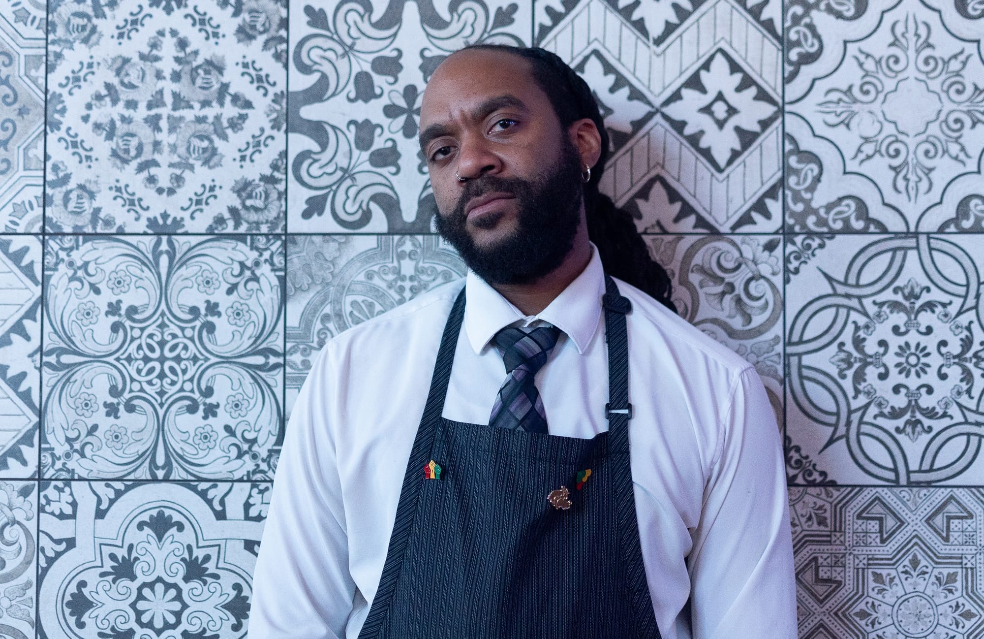 A portrait of a person wearing an apron and leaning against a colorful tiles.