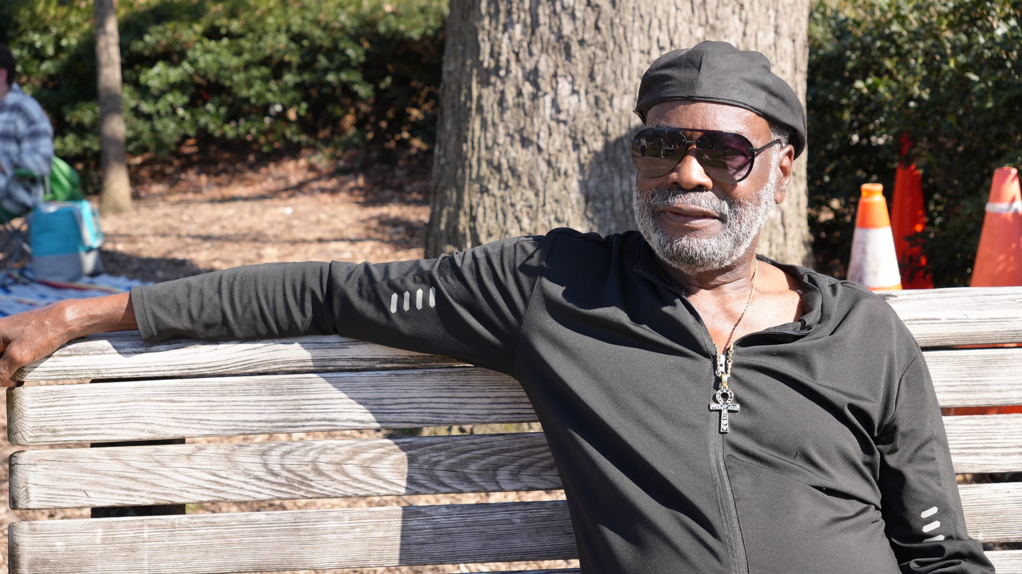 A photo of Mike Gross sitting on a bench. He is wearing a black cap, sunglasses, and a black jacket.
