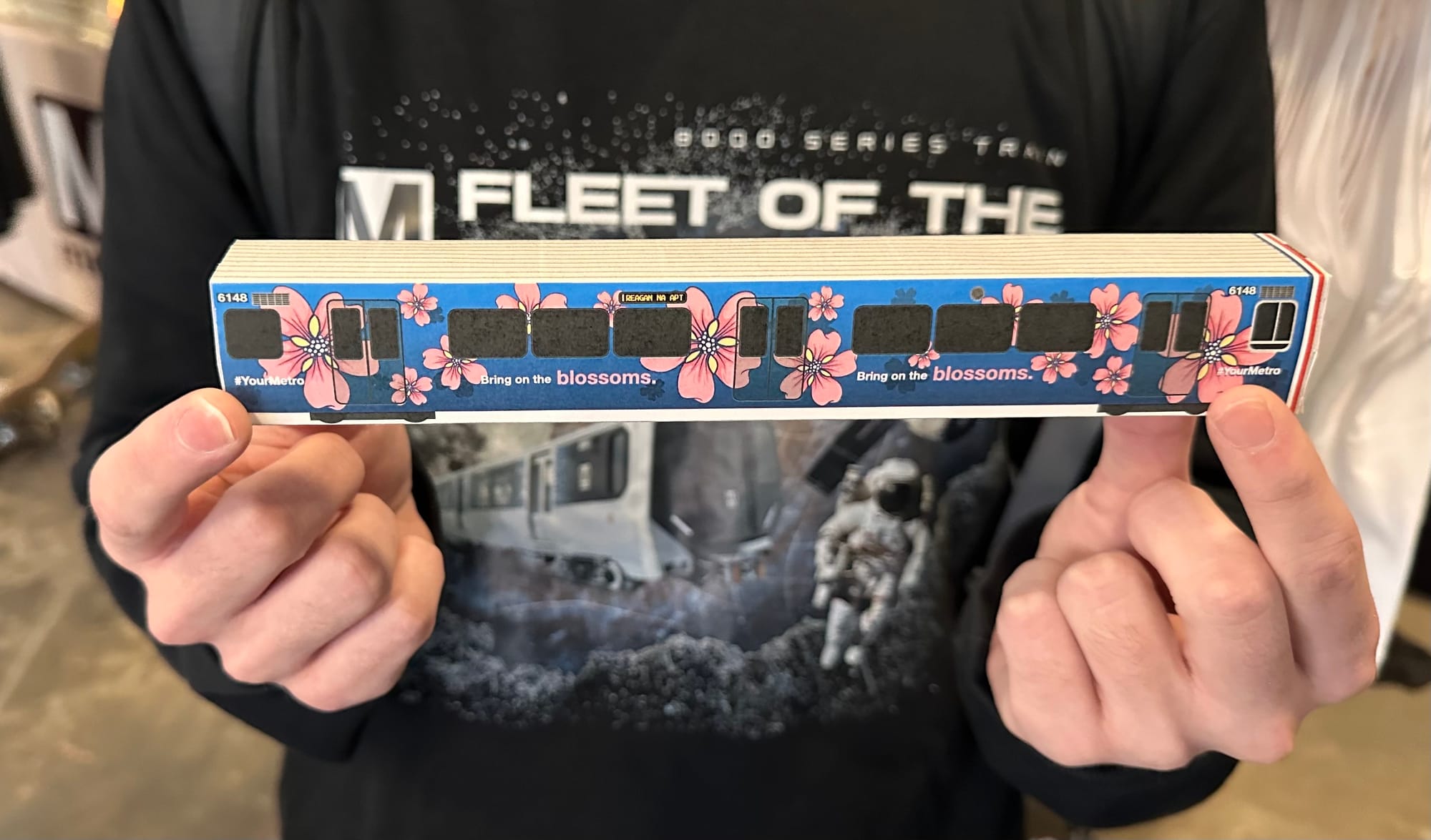 A closeup of two hands holding a paper model of a cherry-blossom themed Metro train 