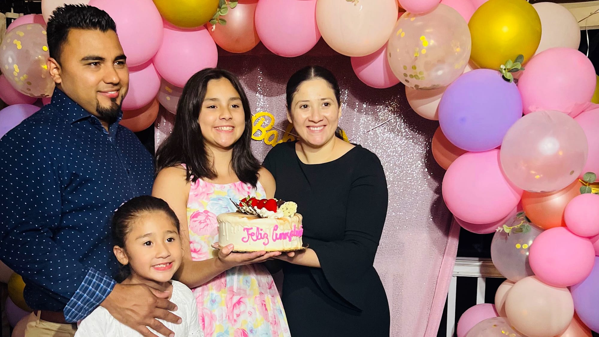 A family of four holds a birthday cake and is surrounded by pink balloons.