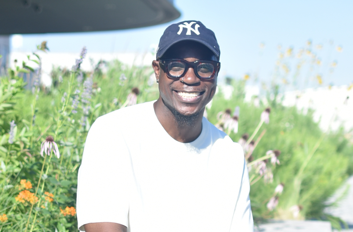 Adewale Agunbiade-Maye sitting infront of flowers growing on the roof of MLK libaray. He's wearing a white shirt, yankees cap and glasses smiling at the camera. 