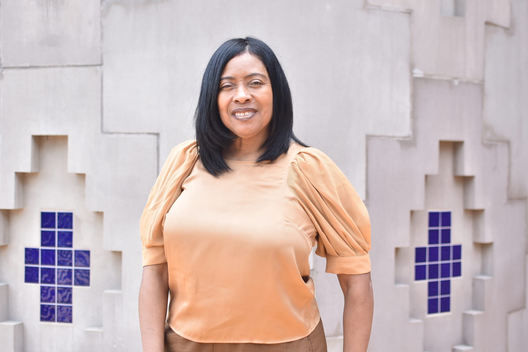 A portrait of Jennifer Morris in front of a white stone wall with blue tile mosaics at the Anacostia Community Museum
