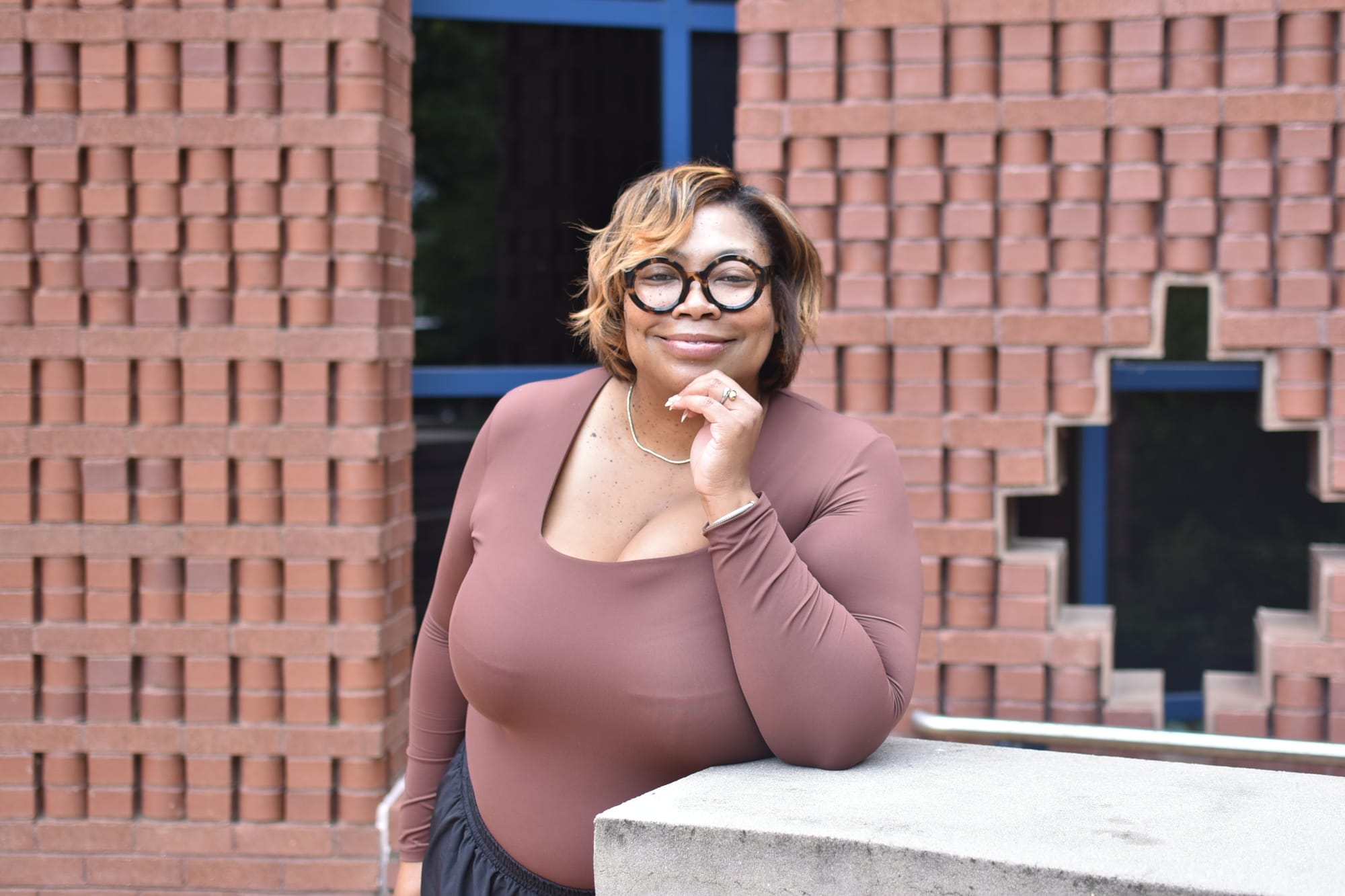 A portrait of Tiffany Penn wearing a brown shirt and glasses, in front of a brick wall at the Anacostia Community Musem