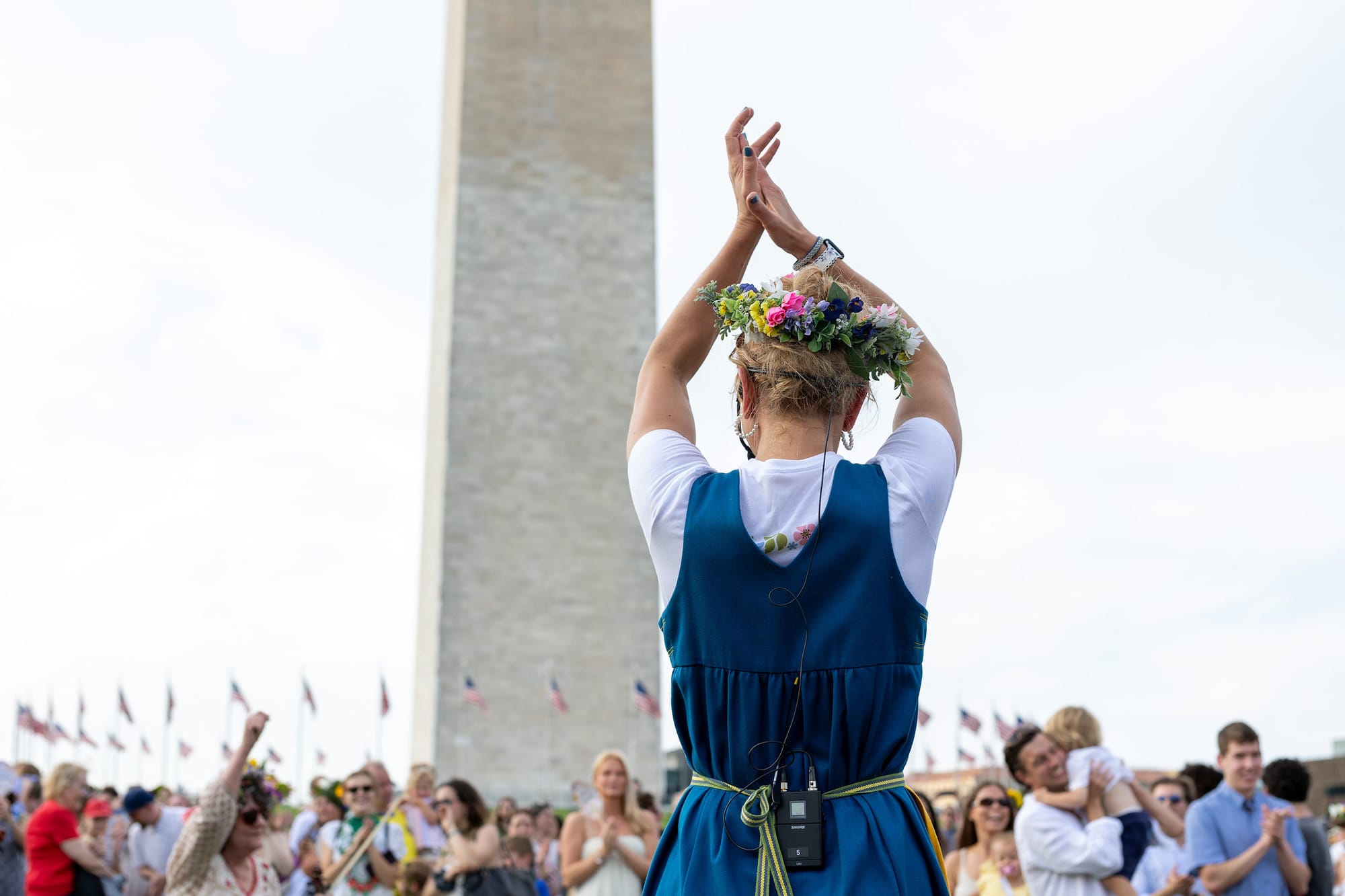 A woman in traditional Swedish clothing and a flower crown stands in front of a crowd at the base of the Washington Monument.