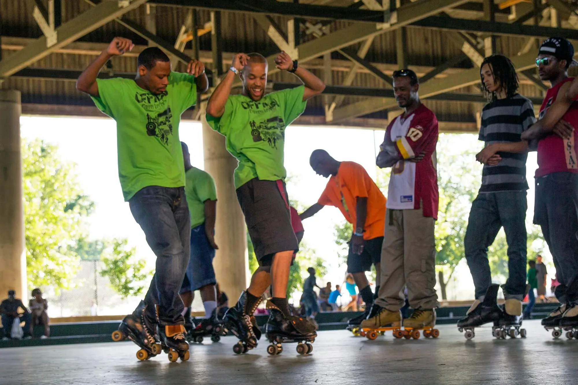 Two people in green shirts do synchronized moves on roller skates while other people in roller skates watch.