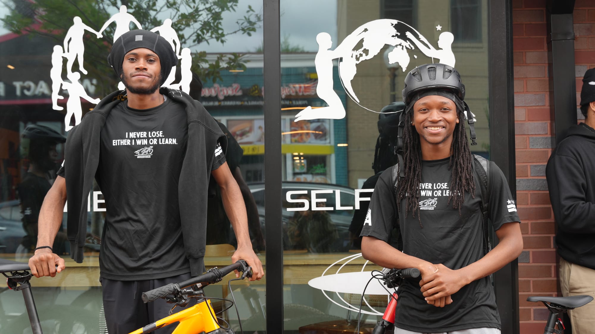 Photo of two students wearing helmets and standing with their bikes in front of a glass and brick storefront.