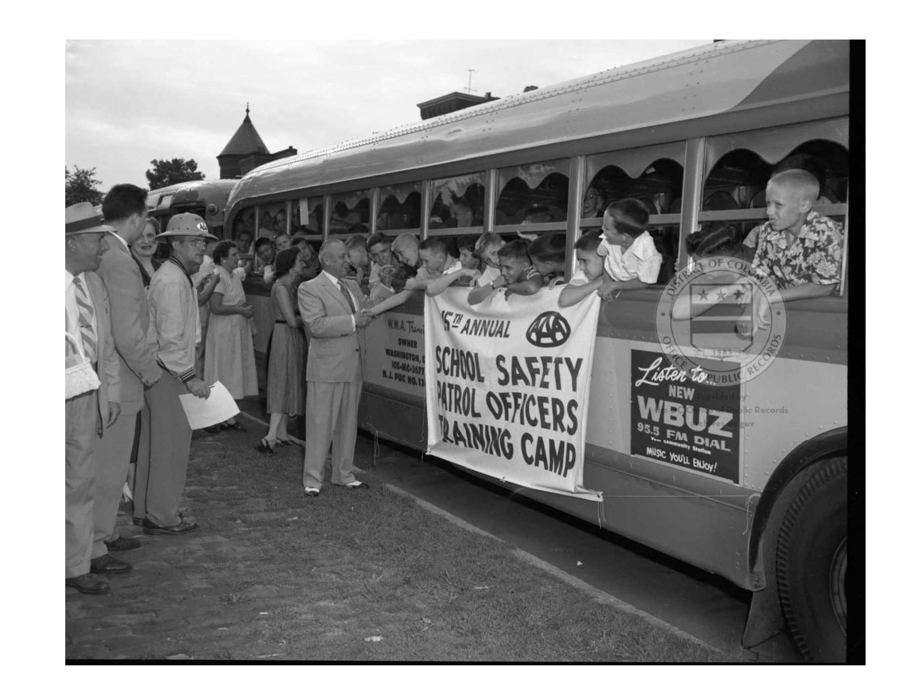 Photo showing a bus with white children leaning out the window. A banner reads "school safety patrol officers training camp"