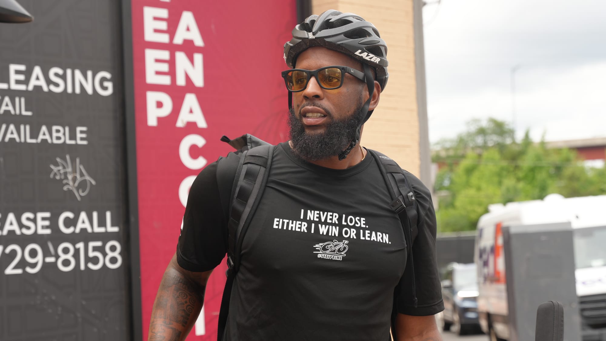 Photo of Clark outside, wearing a backpack, helmet, sunglasses, and a t-shirt that says "I never lose. Either I win or learn."