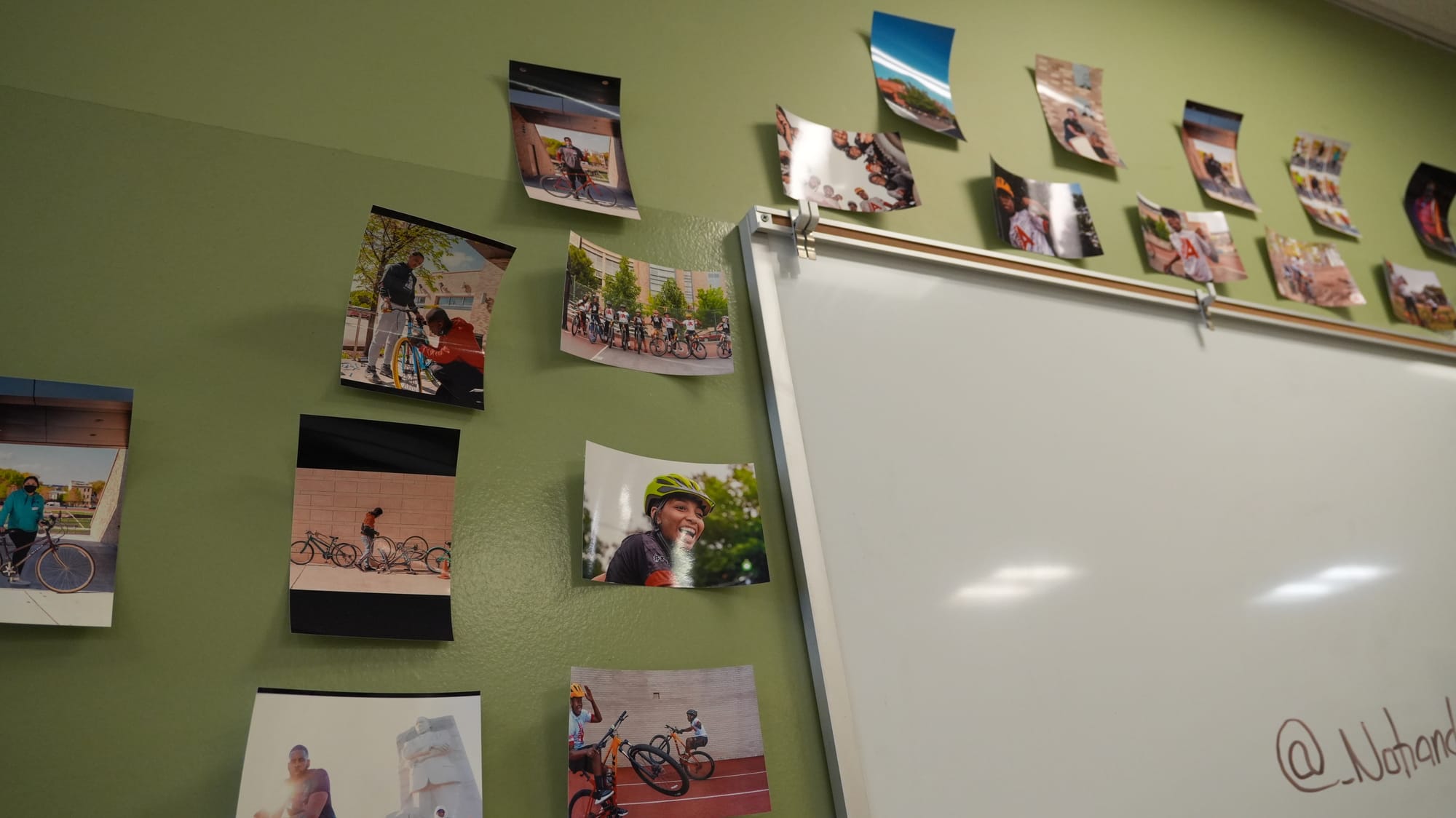 Photo showing a whiteboard on a green wall, with many printed photos stuck to the wall surrounding the board.
