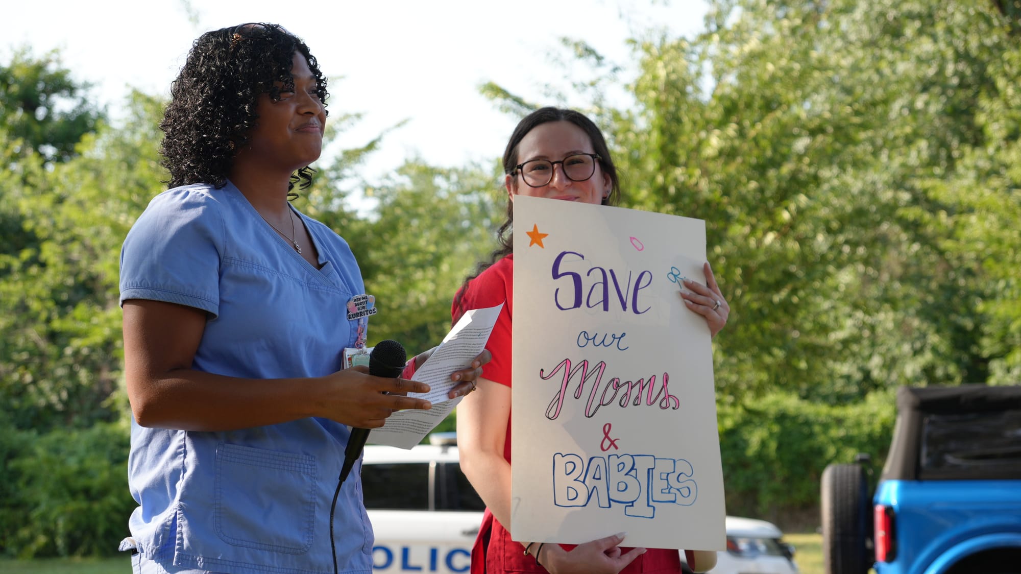 One person in scrubs holding a microphone and another person holding a sign that reads: "Save our moms and babies."