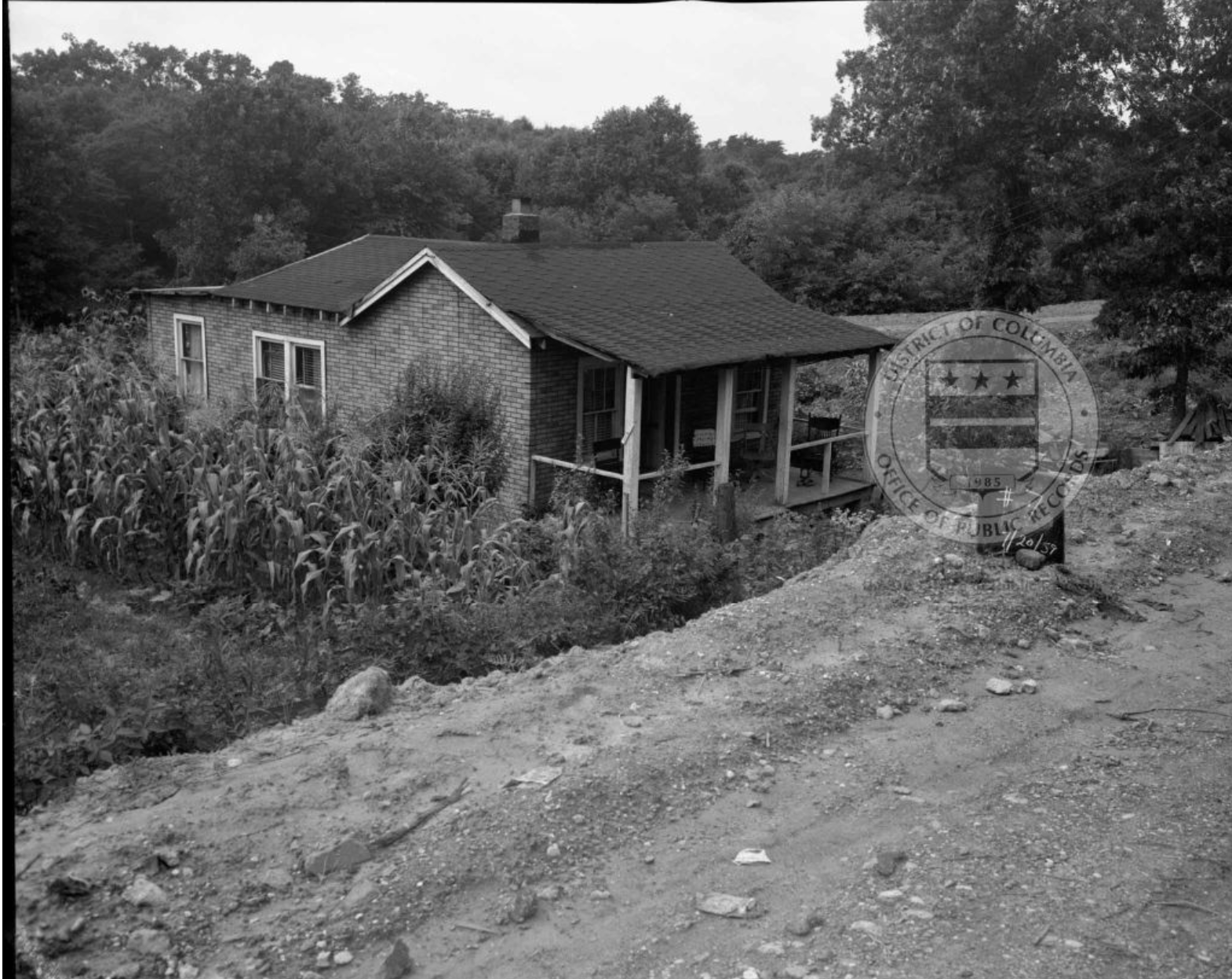 Photo showing a farm-style house with a front porch, set below what appears to be a dirt road.