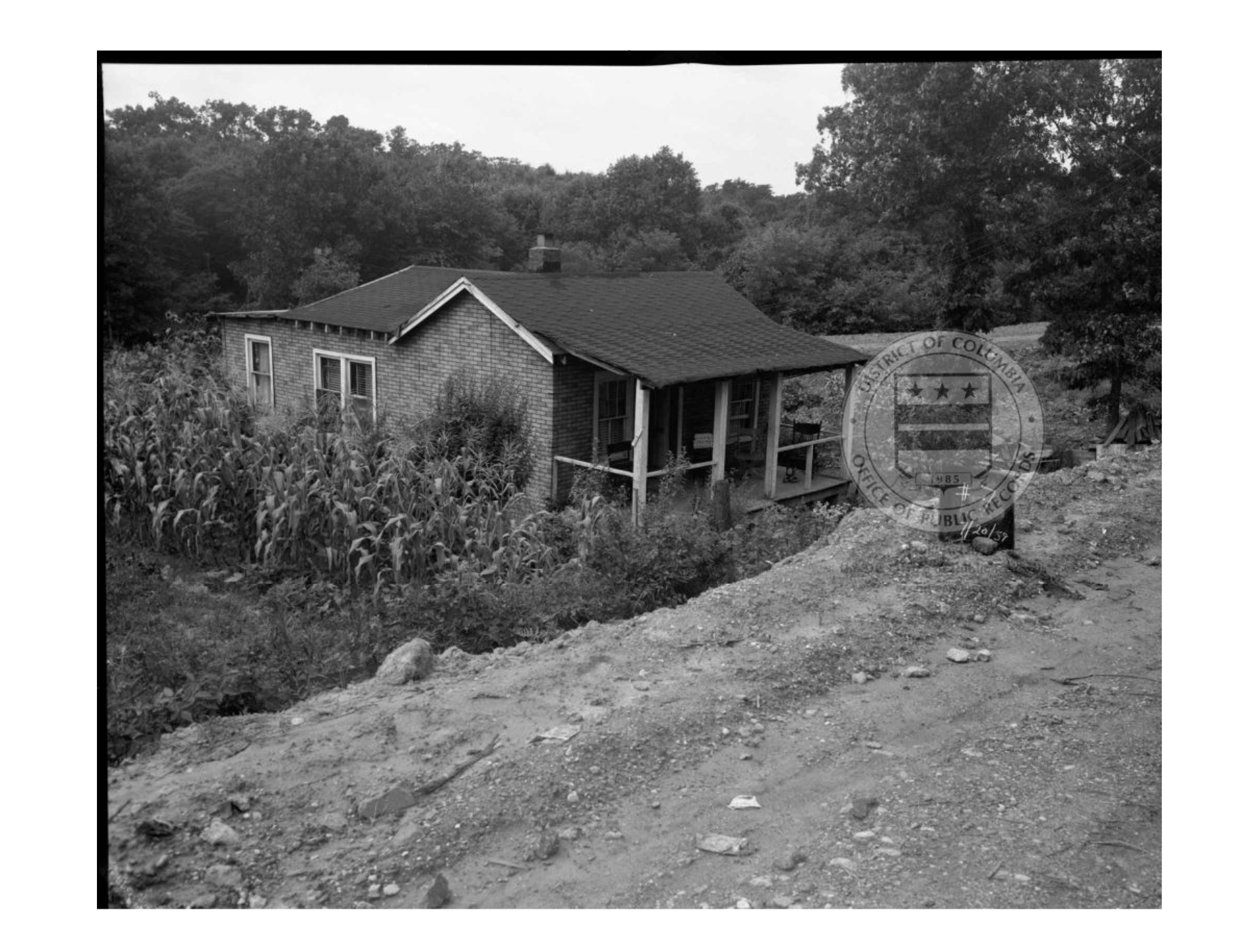 Photo showing a farm-style house with a front porch, set below what appears to be a dirt road.