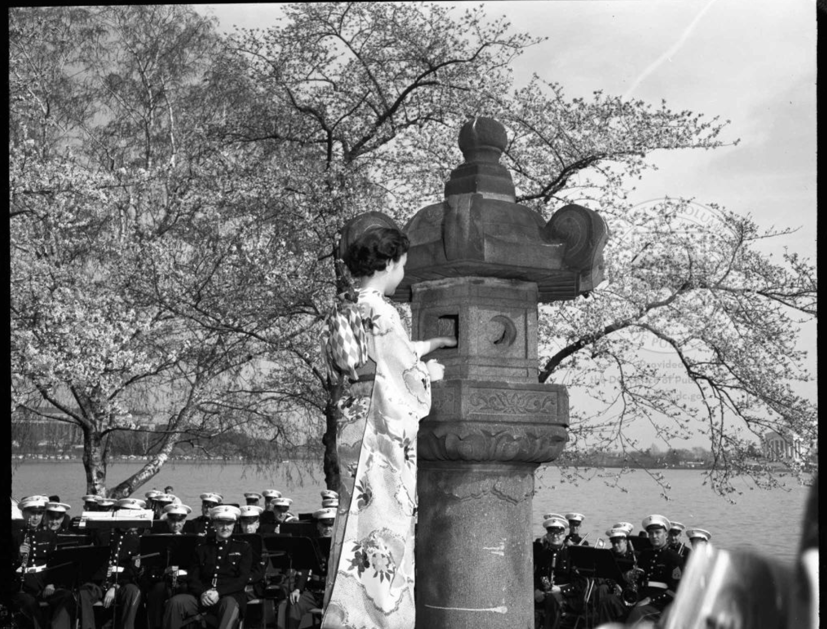 Black and white photo showing a woman in traditional Japanese dress, lighting the lantern. Cherry blossoms and uniformed military members are visible in the background.