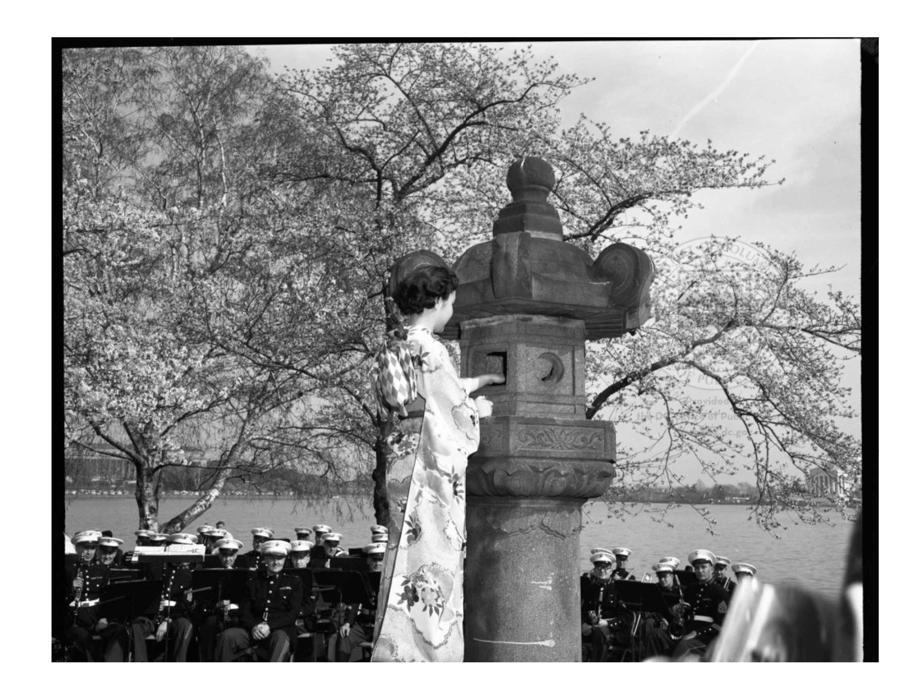 Black and white photo showing a woman in traditional Japanese dress, lighting the lantern. Cherry blossoms and uniformed military members are visible in the background.