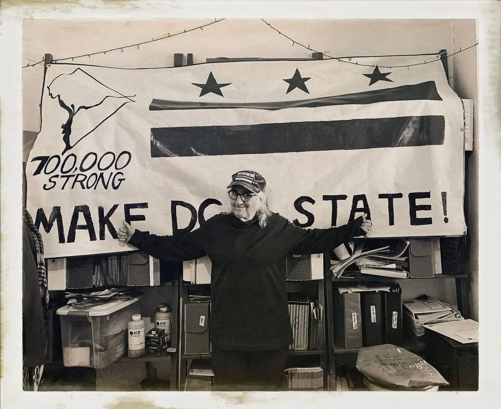 A black and white photograph of Nancy Shia in front of a banner with a D.C. flag that reads: "700,000 strong, make DC a state."