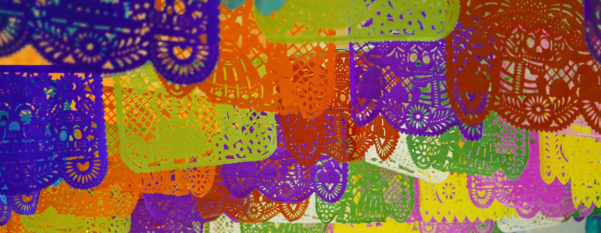 A closeup image of colorful papel picado: Blue, purple, yellow, orange, red, pink, and white sheets.