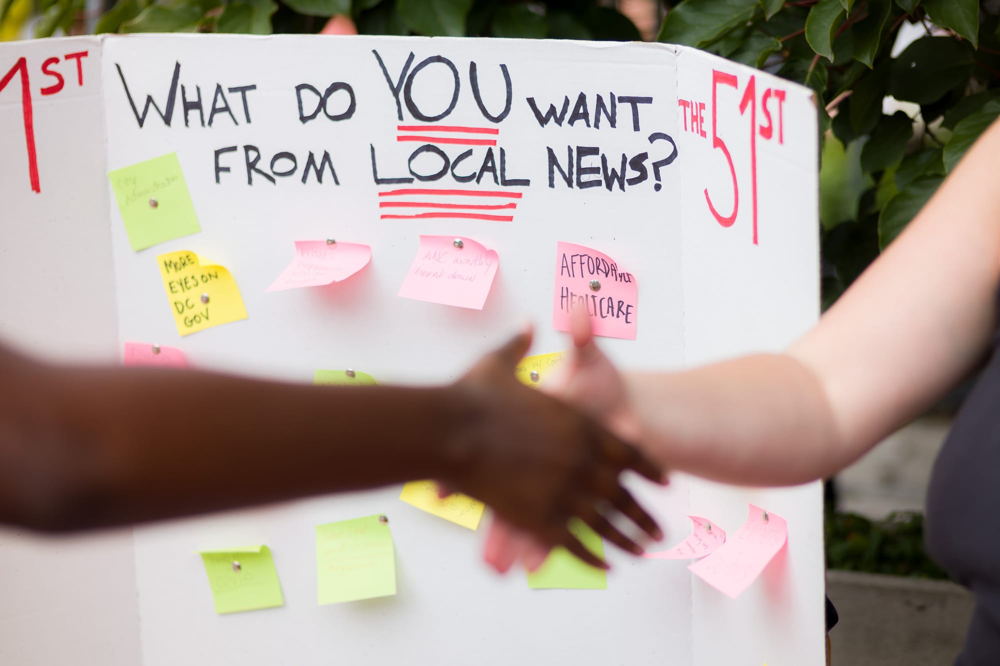 Two people shake hands in front of a poster that reads "What do YOU want from local news?"