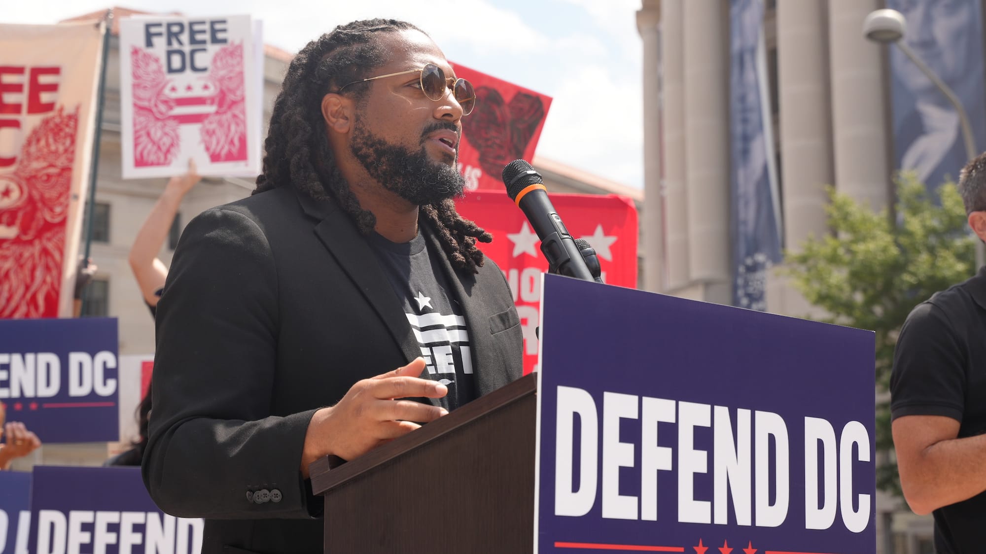 A person speaks at a podium with a sign that says "Defend DC."