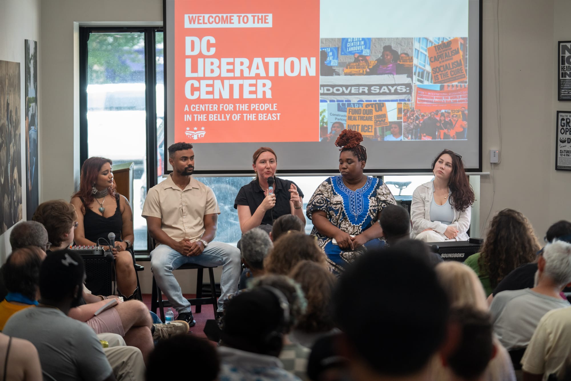 A photo of a group of people speaking to a crowd, with a screen behind them that reads "Welcome to the DC Liberation Center, a center for the people in the belly of the beast."