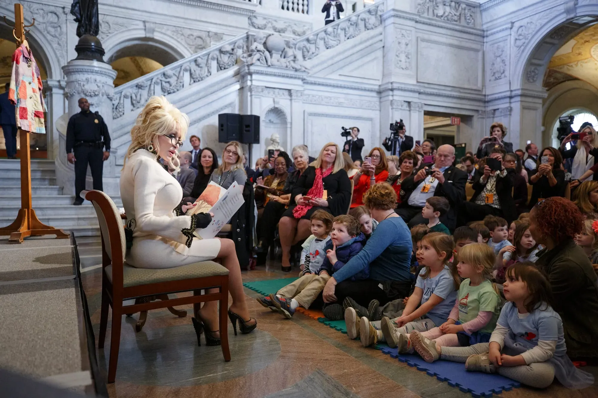 A photo of Dolly Parton wearing white sitting in a chair holding a book and reading to kids.