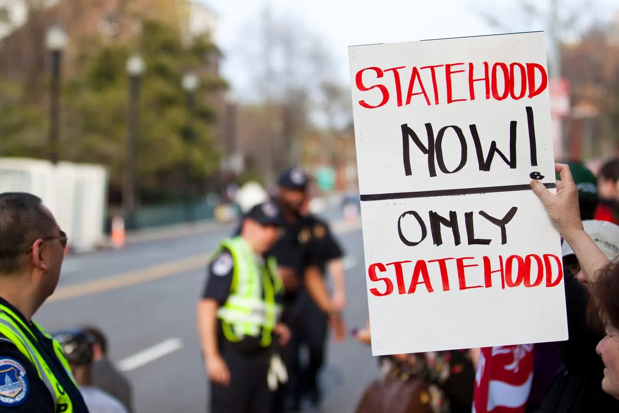 Someone holding a protest sign that reads: "Statehood now! Only Statehood" with police in the background.