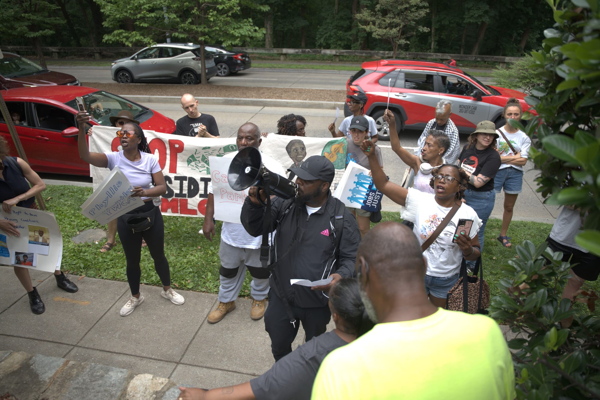 A crowd of tenants are gathered, one with a bullhorn, and many with signs and phones they're using to record.