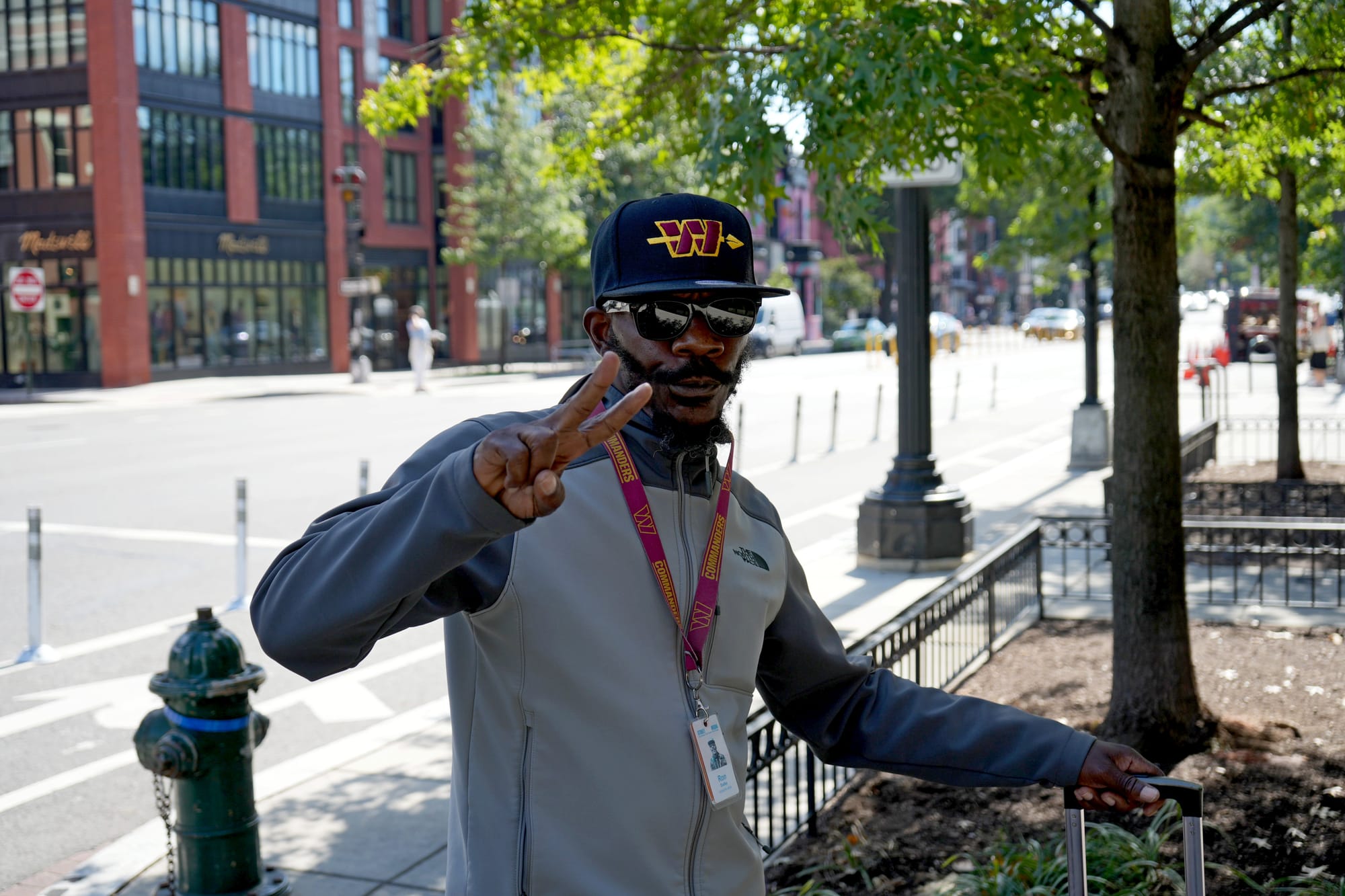 Photo showing a man wearing a Washington Commanders hat and lanyard, attached to his Street Sense Media badge, displaying a "peace" sign with one hand and resting his other hand on a suitcase handle.