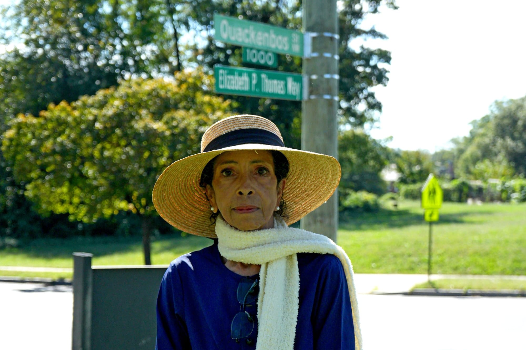 Photo showing a woman staring directly into the camera, wearing a wide-brimmed hat, a scarf, a blue dress, and glasses. The street signs behind her read "Quackenbox; 1000; and Elizabeth P. Thompson Way."