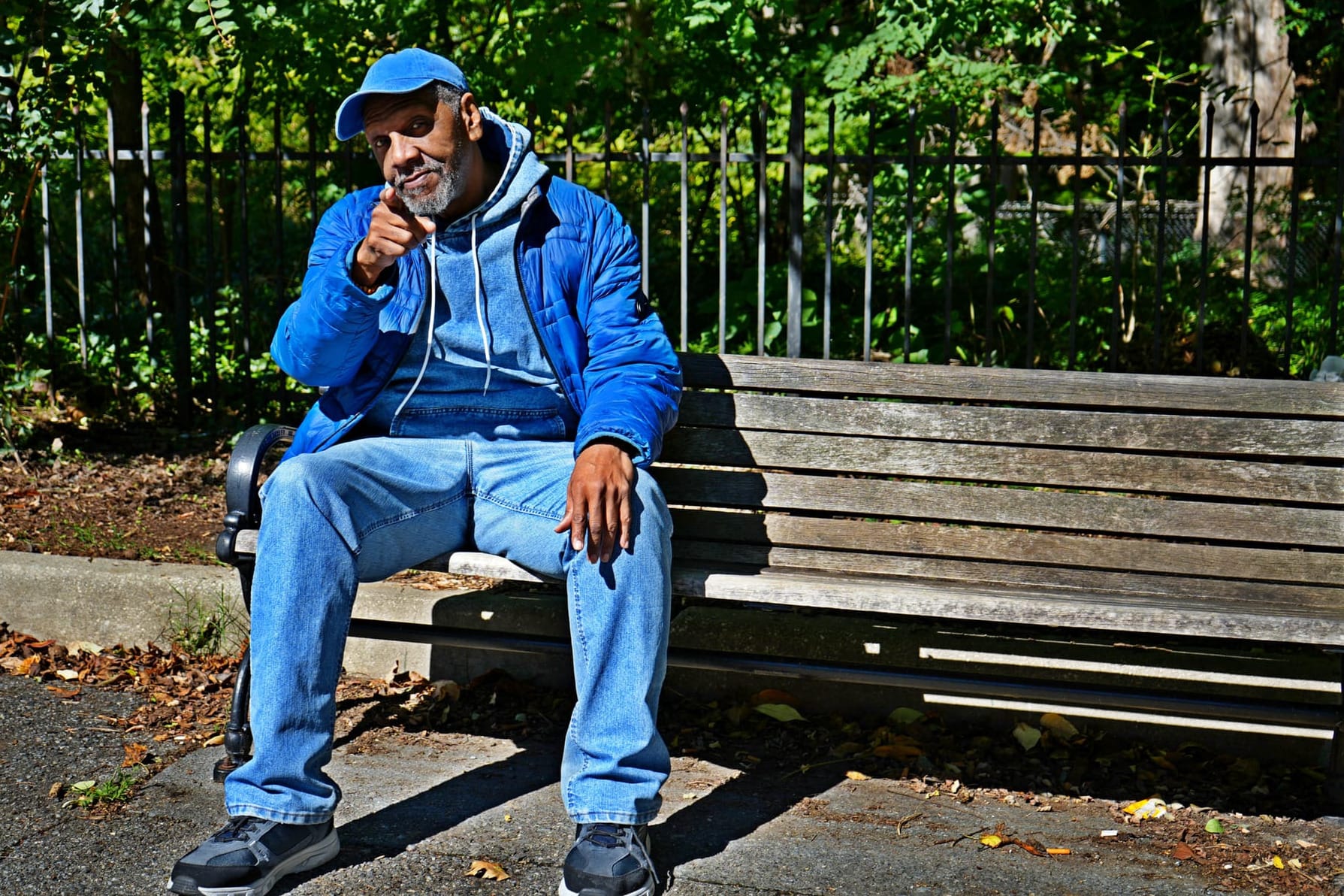 Photo of a man sitting on a park bench, looking directly at the camera with his head cocked to the side and pointing at the viewer.