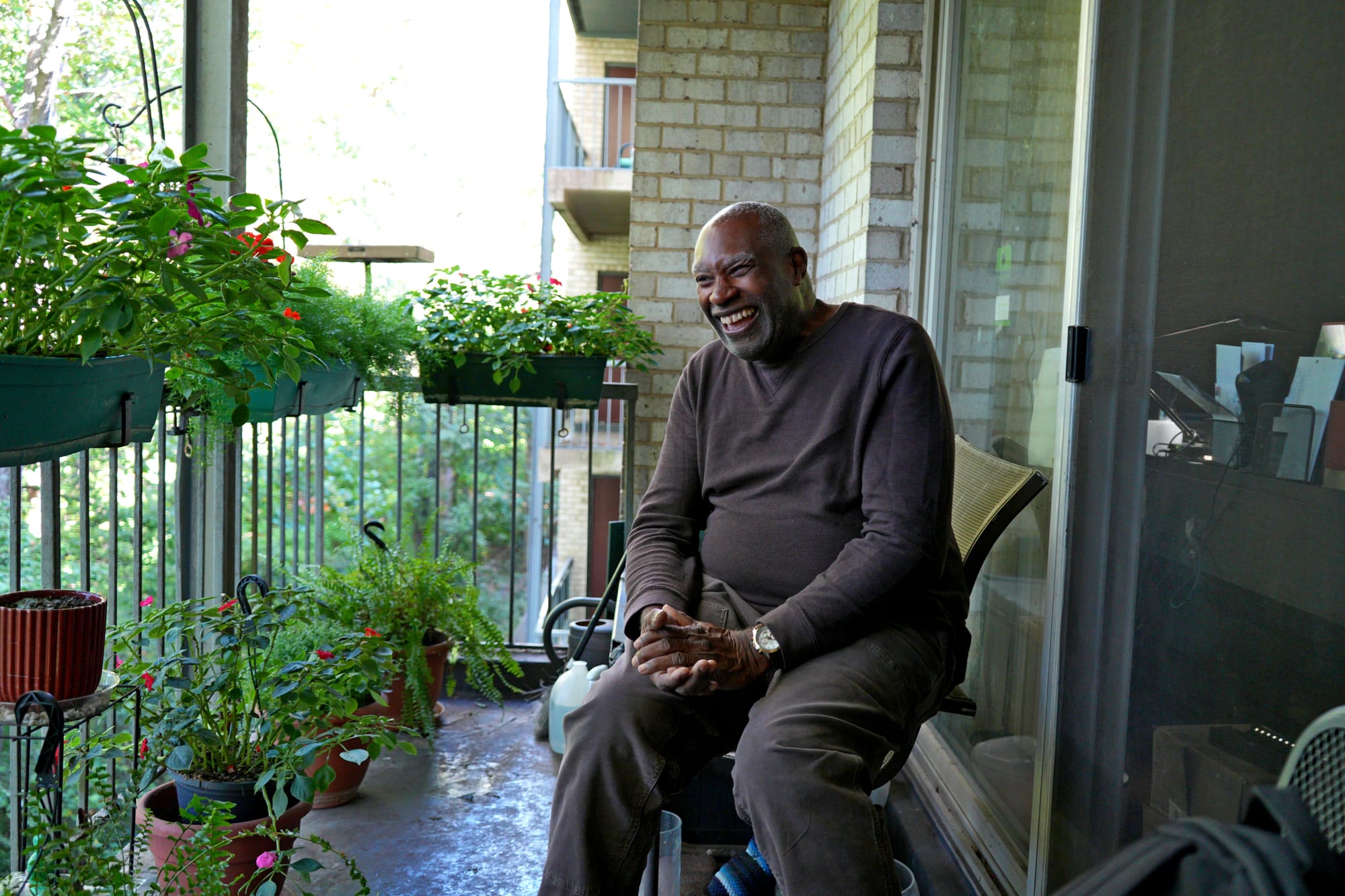 Photo showing a man seated on a porch, laughing, surrounded by potted plants.