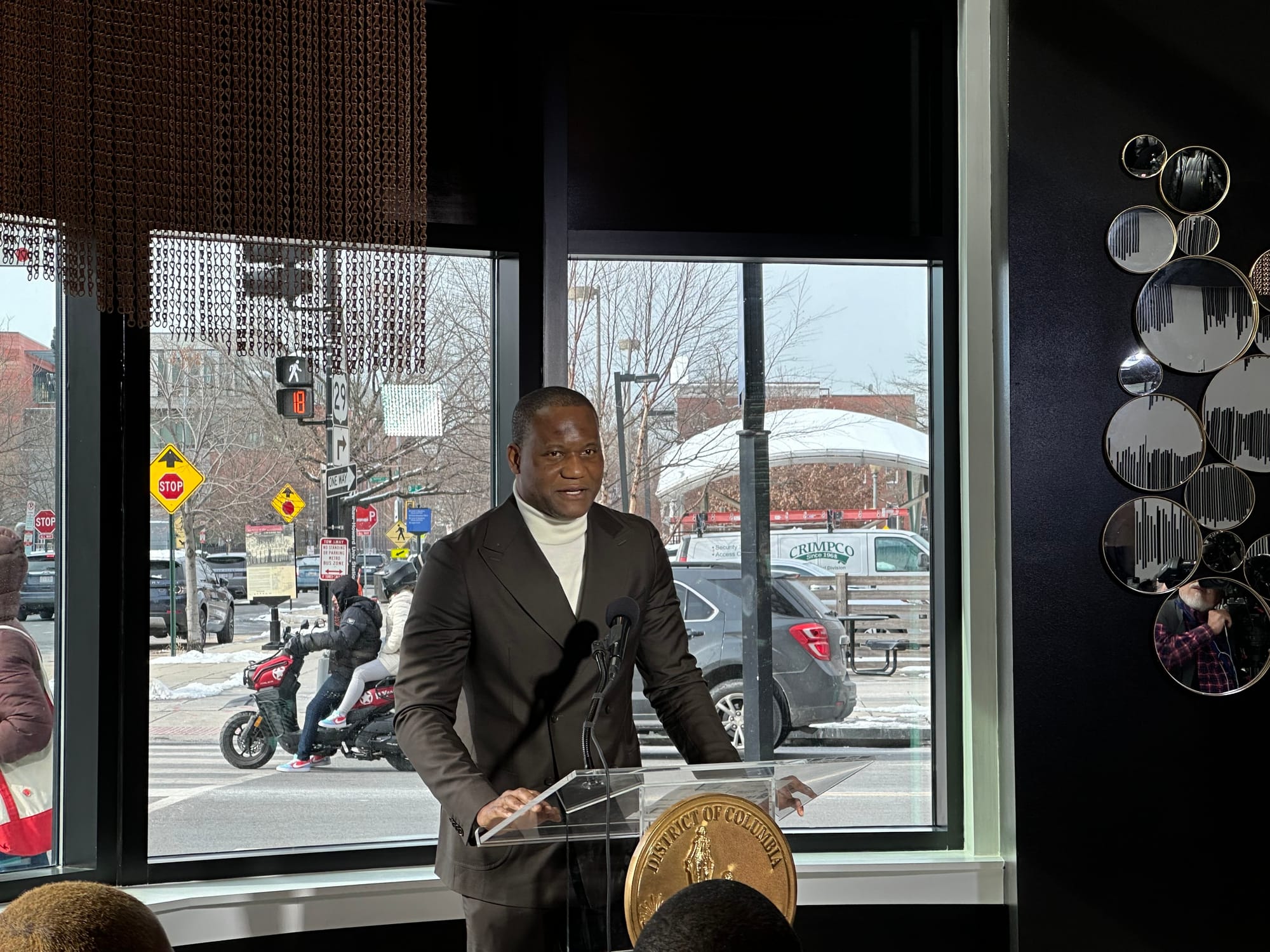Developer Buwa Binitie in front of a podium with a microphone with a window looking onto the street behind him.