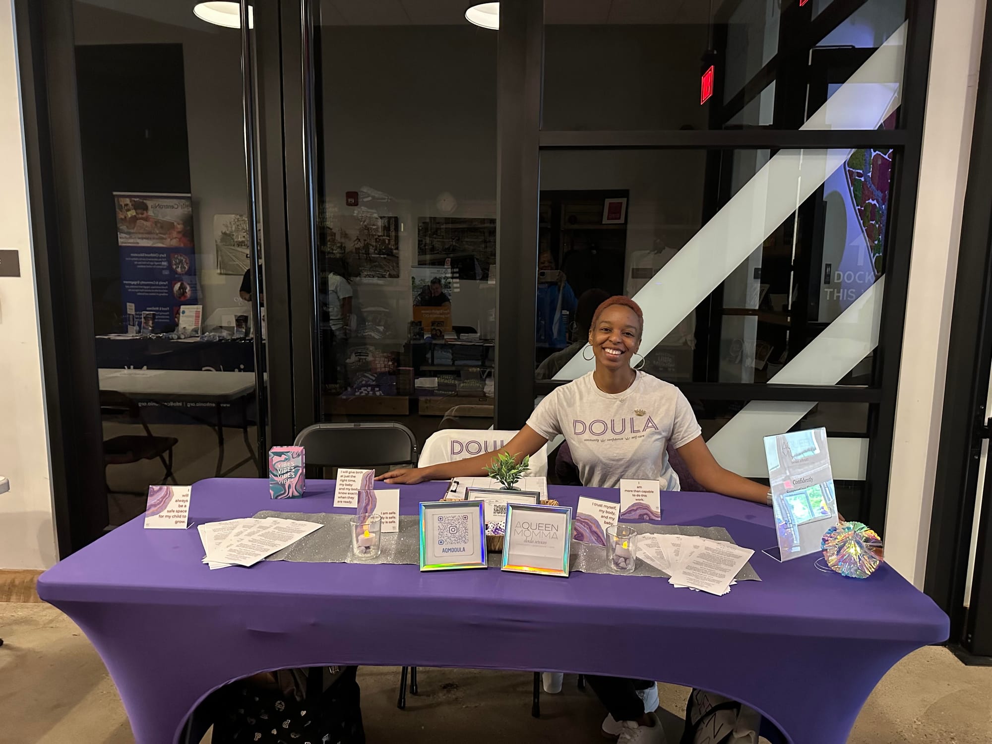 A woman wearing a shirt that says "doula" sitting at a table covered in purple table cloth.