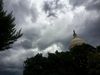 An image of the top of the Capitol dome partially obscured by trees and with a stormy sky in the background.