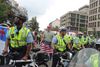 A photograph of a swarm of bicycle cops in yellow vests with a man in the middle wearing a MAGA hat and holding an American flag.