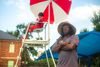 an wearing hat with arms folded standing next to lifeguard stand at pool.