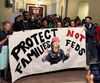 A group of protesters holds up a large banner that reads "Protect families not feds" and shows Muriel Bowser wearing an ICE vest. 