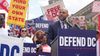 A photo of Robert White speaking at a podium, with people behind him holding up various signs.