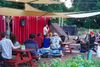 A person performs at an outdoor stage, to an audience at picnic tables and in wooden chairs.