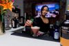 A woman stands behind a bar placing a cherry garnish into reddish non-alcoholic cocktail.