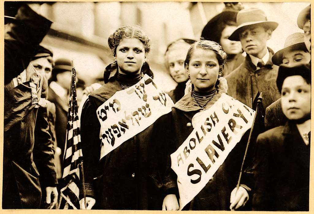 A sepia of two girls in a crowd wearing banners with the slogan "ABOLISH CHILD SLAVERY!!" in English and Yiddish. 