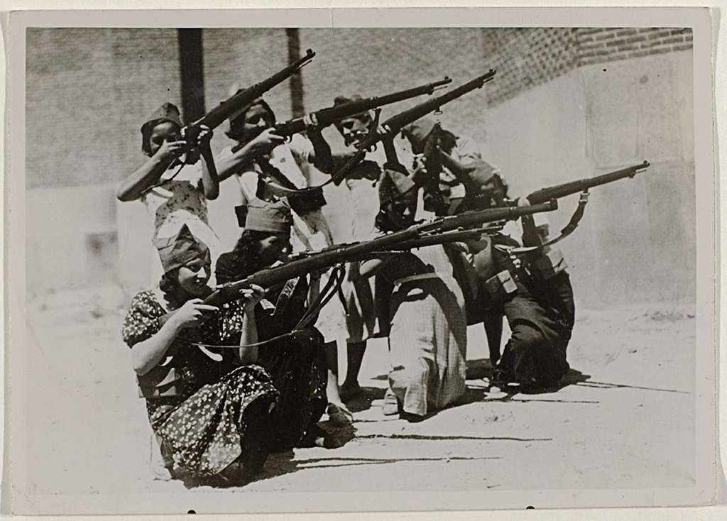 A group of 8 women, 4 crouching, 4 standing, aim their rifles into the distance.